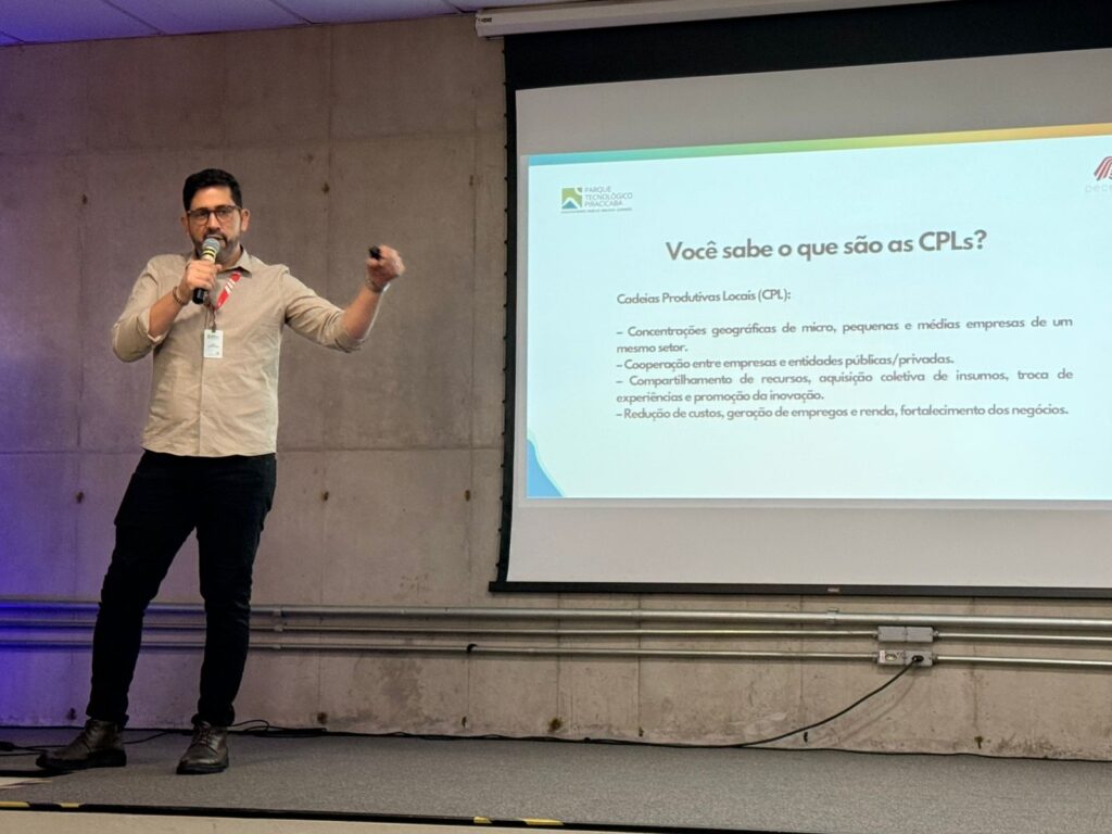 Pedro Chamochumbi detalhou o funcionamento do programa, destacando que as CPLs reúnem empresas e o poder público.jpg descrever A imagem mostra um homem apresentando uma palestra ou workshop em um ambiente interno, possivelmente uma sala de conferências ou auditório. Ele está em pé, segurando um microfone na mão direita e fazendo um gesto com a mão esquerda, indicando que está explicando algo. O homem usa camisa clara de manga comprida dobrada, calça escura, botas e um crachá pendurado no pescoço. Ao fundo, há um grande telão de projeção exibindo um slide com o título "Você sabe o que são as CPLs?" e o logotipo do "Parque Tecnológico Piracicaba" no canto superior esquerdo. O slide explica o conceito de "Cadeias Produtivas Locais (CPL)" em português, destacando pontos como a concentração geográfica de empresas de pequeno e médio porte, cooperação entre empresas e entidades, compartilhamento de recursos, inovação, redução de custos e geração de empregos. A parede atrás do apresentador é de concreto com uma textura visível, e há algumas instalações elétricas aparentes próximas ao chão. A iluminação é clara e uniforme, sugerindo luz artificial de teto. O ângulo da foto é ligeiramente de baixo para cima, como se o fotógrafo estivesse sentado na plateia.