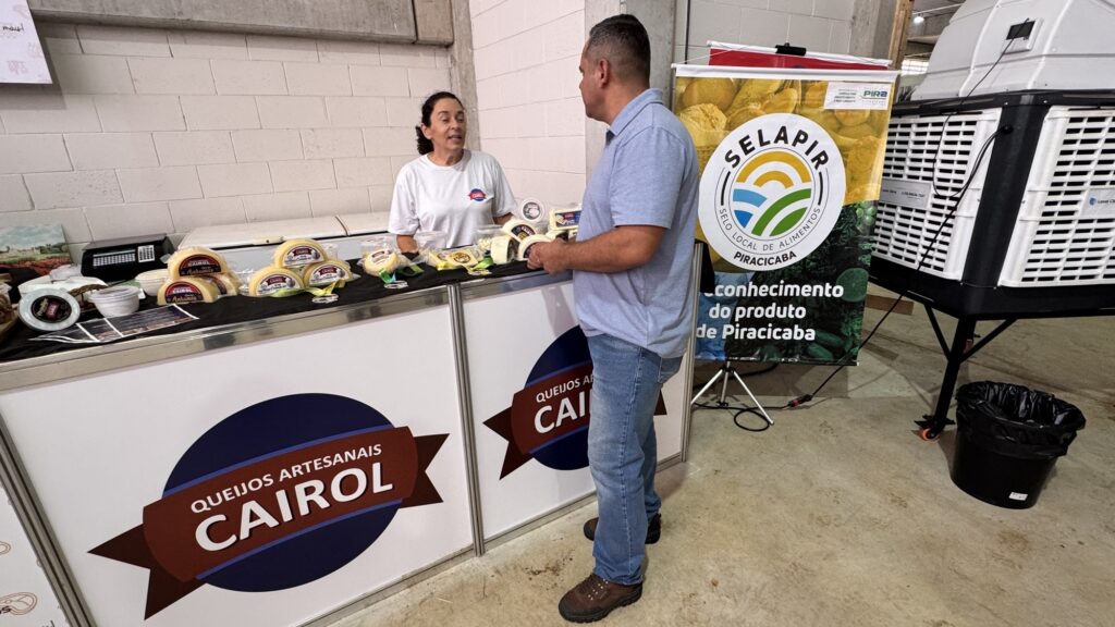 A imagem mostra uma barraca de queijos artesanais chamada "Queijos Artesanais Cairol", possivelmente em uma feira ou mercado local. Uma mulher, vestindo camiseta branca e com cabelo preso, está atrás do balcão, aparentemente atendendo ou conversando com um homem que está do outro lado do balcão. O homem veste uma camisa azul clara, calça jeans e botas marrons. Sobre o balcão, há vários pedaços e embalagens de queijo expostos para venda, todos etiquetados com a marca "Cairol". Ao lado da barraca, há um banner vertical com o logotipo "SELAPIR", que é o selo local de alimentos de Piracicaba, indicando que o produto é típico ou certificado da região. O ambiente parece ser interno, com paredes brancas e um equipamento grande ao fundo, possivelmente um refrigerador industrial para manter os queijos frescos. A cena transmite uma atmosfera de comércio local, valorizando produtos artesanais e regionais.