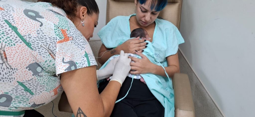 A imagem mostra uma cena em um ambiente hospitalar, provavelmente em uma unidade neonatal. Uma mulher, vestindo um avental hospitalar azul claro com a inscrição "HFC Saúde", está sentada em uma cadeira, segurando um bebê recém-nascido no colo. O bebê é pequeno, com cabelos escuros, e está apoiado no peito da mãe. Ao lado da mulher, há um profissional da saúde, possivelmente uma enfermeira, usando um uniforme branco com estampas de animais (elefantes, girafas e crocodilos). Ela está usando luvas brancas e parece estar realizando algum procedimento ou exame no bebê, demonstrando cuidado e atenção. O ambiente é claro e limpo, com paredes brancas, transmitindo a sensação de um local hospitalar bem organizado. A interação entre a mãe, o bebê e a profissional da saúde sugere um momento de cuidado e acompanhamento médico no pós-parto.