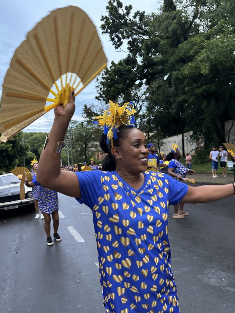 A imagem mostra uma mulher negra sorridente participando de um desfile ou festa de rua. Ela veste uma camiseta azul decorada com corações dourados e usa uma tiara elaborada feita de penas amarelas e azuis, além de lantejoulas douradas. Na mão direita, ela segura um grande leque amarelo aberto, que está em movimento, sugerindo que ela está dançando ou se movimentando. Ela também usa uma pulseira larga prateada no braço direito. Ao fundo, outras pessoas também vestem roupas azuis e amarelas, algumas segurando leques semelhantes, e parece ser um evento ao ar livre com árvores verdes ao redor. Algumas pessoas observam o desfile na calçada. A cena é iluminada por luz natural em um dia nublado. A composição transmite alegria e energia festiva.