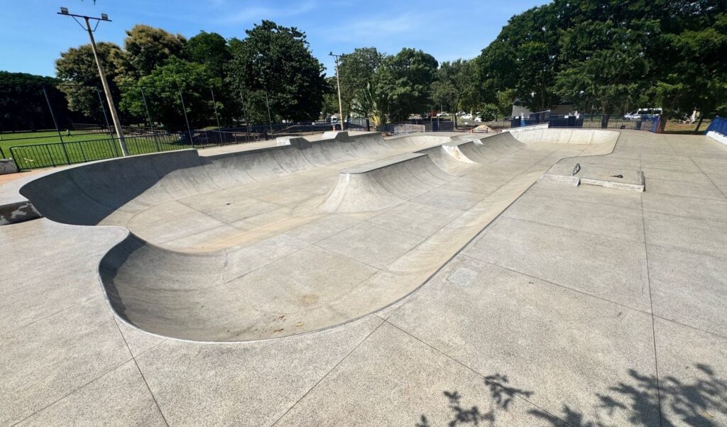 A imagem mostra um skatepark ao ar livre, com uma grande pista de concreto em formato de bowl, ideal para a prática de skate. A pista possui várias transições e elevações, incluindo uma espécie de lombada central. As bordas do bowl são revestidas com um metal para facilitar as manobras. O local está cercado por árvores verdes e há uma grade metálica visível à direita, possivelmente para segurança. O céu está limpo e azul, indicando um dia ensolarado. Não há pessoas na imagem, e o ambiente transmite uma sensação de espaço aberto e bem cuidado, ideal para atividades recreativas.