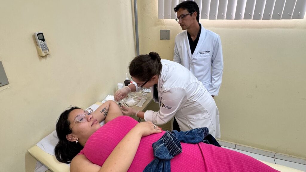 A imagem mostra um ambiente clínico, provavelmente uma sala de atendimento médico. No centro, uma paciente está deitada em uma maca, vestindo uma blusa rosa de mangas compridas e óculos. Ela está com o braço direito levantado, exibindo uma tatuagem, enquanto uma profissional de saúde, vestida com jaleco branco e luvas, aplica uma injeção ou realiza uma coleta de sangue em seu braço. Atrás da profissional, um médico, também de jaleco branco e usando óculos, observa atentamente o procedimento. O ambiente é bem iluminado, com paredes amarelas claras e uma janela com persianas brancas. Ao lado da maca, há uma mesa com diversos materiais médicos, como frascos, algodão e seringas. Na parede, há um controle remoto fixado. No canto inferior da imagem, aparecem informações escritas: "Dr. Martin Eduardo Monclaire", "Ginecologia e Obstetrícia", "São Leopoldo Mandic" e "Elgin", indicando provavelmente o nome do médico, sua especialidade e o local de atendimento. A cena transmite um ambiente profissional e de cuidado médico.