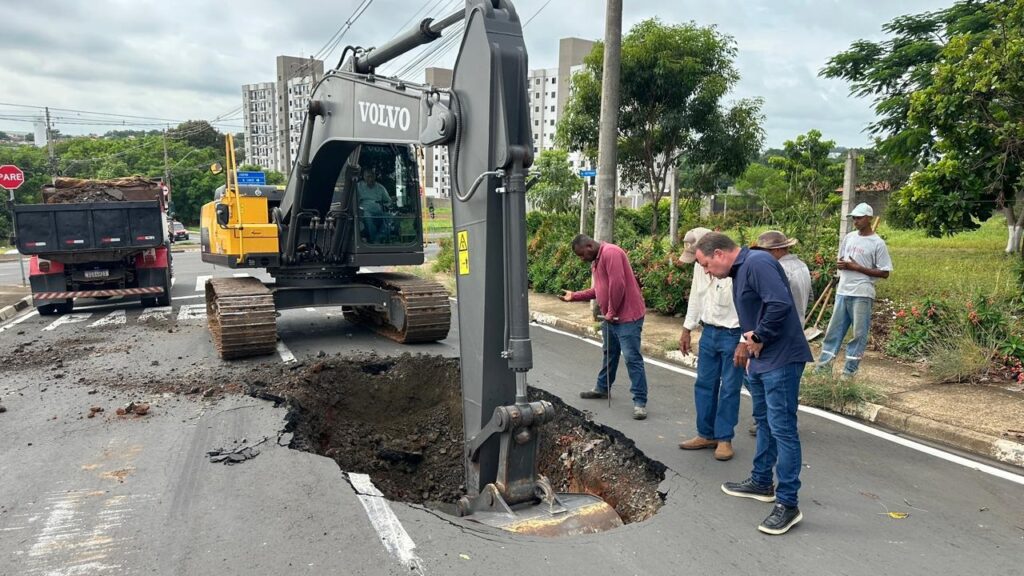 A imagem mostra uma cena de manutenção viária em um dia nublado. No centro, há uma escavadeira amarela da marca Volvo, com o braço abaixado dentro de um grande buraco na pista de asfalto. Ao lado esquerdo da escavadeira, há um caminhão basculante vermelho e preto, parcialmente carregado com entulho. Várias pessoas estão próximas ao buraco, observando ou trabalhando: um homem de camiseta vinho segurando uma ferramenta, outro de camisa azul de manga longa e jeans olhando atentamente para o buraco, além de outras pessoas vestindo roupas informais, algumas com chapéus. Ao fundo, é possível ver prédios residenciais de vários andares, árvores e postes de energia elétrica. No canto da imagem, há uma placa de trânsito vermelha com a palavra "PARE". A cena transmite um momento de reparo ou inspeção na via, com o uso de maquinário pesado e presença de trabalhadores.