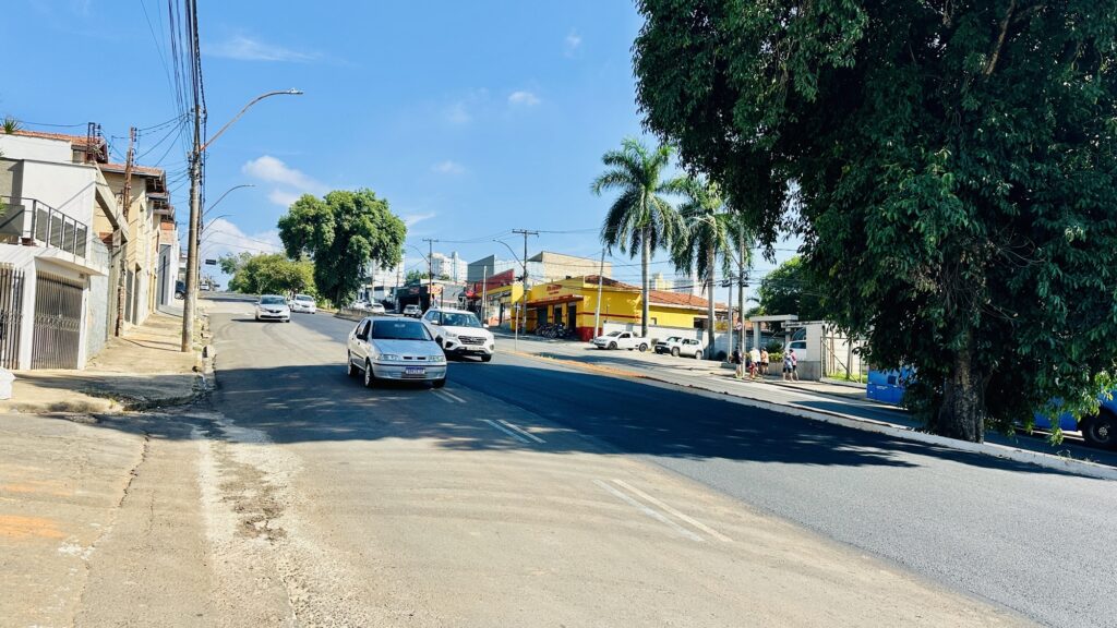 A imagem mostra uma rua asfaltada em uma área urbana, sob um céu azul com poucas nuvens brancas. A rua está levemente inclinada para cima, com vários carros estacionados e em movimento, incluindo um carro prata em primeiro plano com a placa "DAX2E17". À direita, há uma grande árvore com folhagem verde densa que ocupa parte da calçada. No fundo, do lado esquerdo, há um prédio amarelo com a palavra "MOTO" visível na fachada, sugerindo um comércio local. A rua é ladeada por postes de energia com fios cruzando o céu, e algumas pessoas podem ser vistas caminhando na calçada ao longe. A cena transmite um ambiente típico de um bairro residencial e comercial, possivelmente em uma cidade do Brasil, devido ao estilo dos carros, placas e vegetação tropical.
