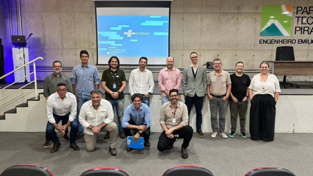 A imagem mostra um grupo de treze pessoas posando para uma foto em um ambiente corporativo, possivelmente uma sala de conferências ou auditório. O grupo está dividido em duas fileiras: nove pessoas estão em pé na parte de trás e quatro estão agachadas na frente. A maioria dos indivíduos é do sexo masculino, com uma mulher posicionada à direita. Eles vestem roupas formais e semi-formais, como camisas sociais, polos, blazers e calças. Ao fundo, há uma parede de concreto com uma grande tela de projeção exibindo um slide azul com o logotipo do SEBRAE (Serviço Brasileiro de Apoio às Micro e Pequenas Empresas). À direita da tela, há um grande letreiro na parede que diz "PARQUE TECNOLÓGICO PIRACICABA" e "ENGENHEIRO EMÍLIO". Do lado esquerdo, observa-se um pequeno palco com um púlpito, um notebook sobre um suporte e uma escada. A iluminação é clara e uniforme, típica de ambientes internos corporativos. A atmosfera da foto é profissional, indicando que o grupo pode estar participando de um evento, treinamento ou reunião