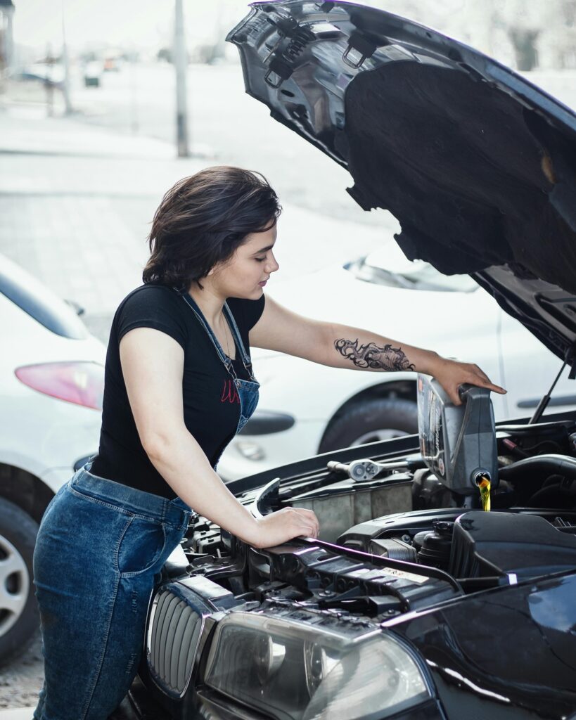A imagem mostra uma mulher jovem realizando a manutenção de um carro, especificamente adicionando óleo ao motor. Ela está vestindo uma camiseta preta e um macacão jeans, e possui uma tatuagem grande e escura no braço esquerdo. O carro é um BMW preto com o capô aberto, e a mulher está concentrada enquanto despeja o óleo de um recipiente cinza. Ao redor, é possível ver outras pessoas e carros, sugerindo que a cena acontece em um ambiente urbano ao ar livre durante o dia. A luz natural destaca os detalhes da cena, e há uma chave de soquete apoiada próxima ao motor, indicando que ela pode estar realizando uma manutenção mais completa no veículo.