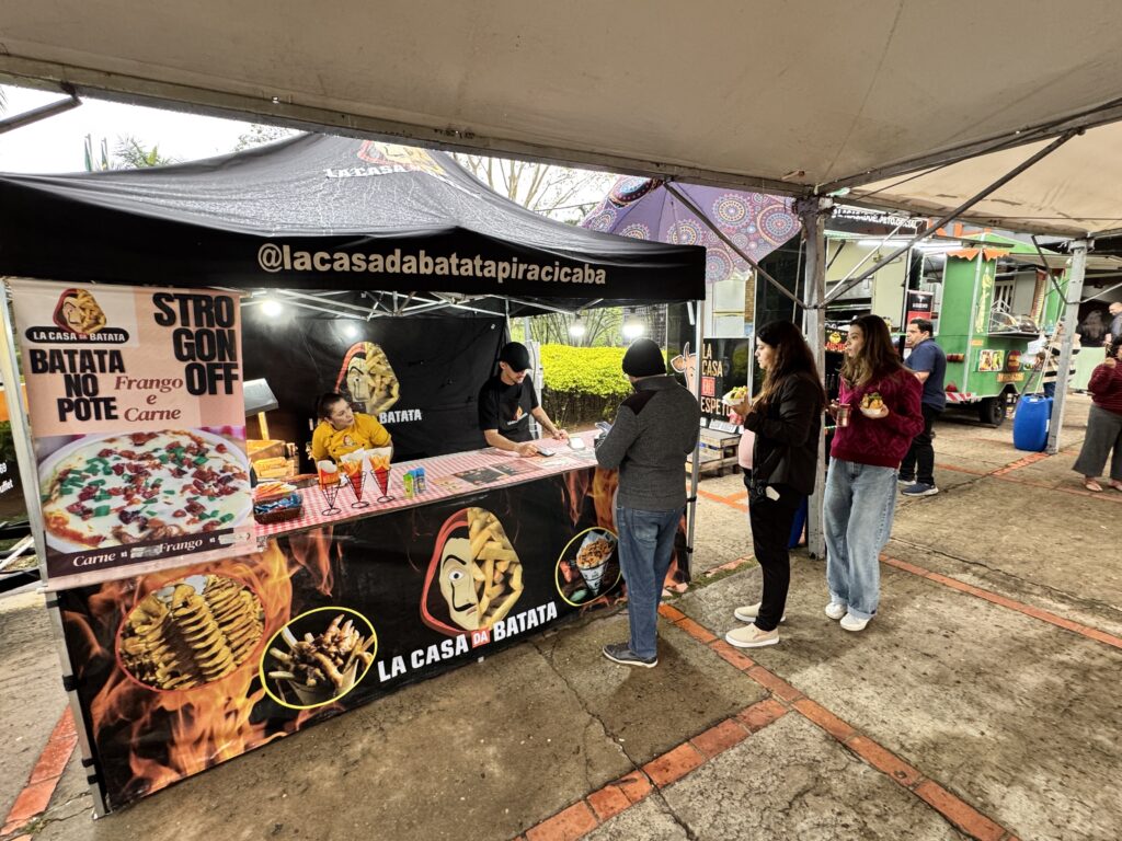 A imagem mostra uma barraca de comida chamada "LA CASA DA BATATA" em um evento ao ar livre, possivelmente um festival de comida. A barraca está montada sob uma tenda preta e apresenta uma logo que lembra a série "La Casa de Papel", com uma máscara de Salvador Dalí estilizada com batatas fritas no lugar do cabelo e capuz. Na barraca, há dois funcionários atendendo os clientes: uma mulher vestindo uma camiseta amarela e um homem com boné e camiseta preta. Na frente da barraca, um grupo de pessoas está na fila, esperando para fazer seus pedidos. Entre os clientes, há uma mulher com uma blusa vermelha e calça jeans larga, segurando um prato com comida. No cardápio da barraca, há opções como "Batata no Pote" com frango e carne, além de strogonoff nas versões carne e frango. A barraca parece especializada em pratos com batata, e o ambiente é descontraído, com outras barracas e food trucks ao fundo, sob uma cobertura branca. O chão é de piso intertravado com detalhes em vermelho, e o clima aparenta ser de dia nublado.