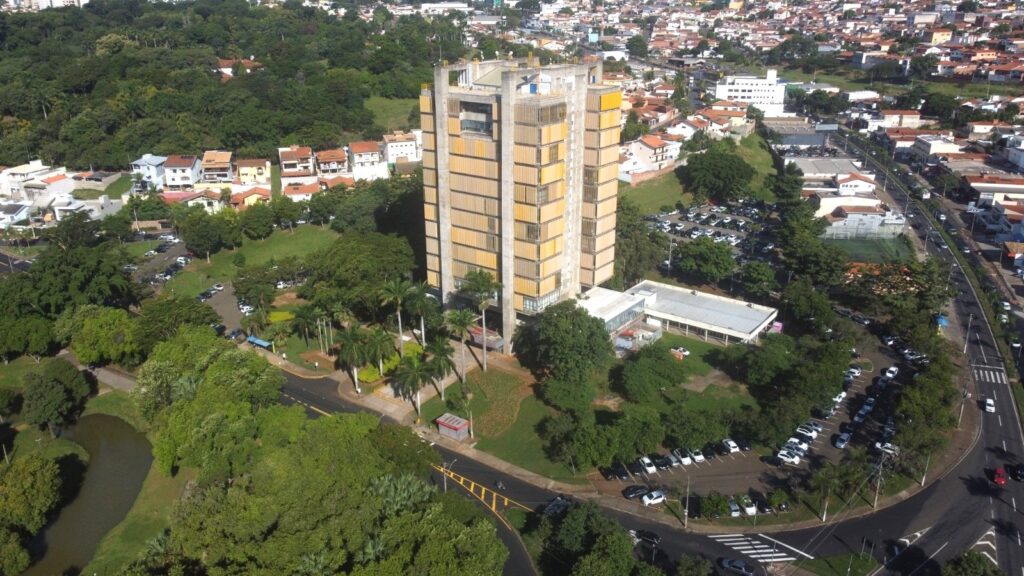 A imagem mostra uma vista aérea de um edifício alto e marcante, estilo brutalista, com painéis amarelos que se destacam em sua fachada. O prédio tem uma estrutura de concreto aparente e parece ser um ponto central da área, possivelmente um edifício público ou institucional. Ao redor do edifício principal, há áreas verdes bem cuidadas, com muitas árvores, além de estacionamentos com vários carros. Próximo ao prédio, há construções menores, de cor clara, que parecem ser anexos ou outras instalações relacionadas. A área ao redor mistura elementos urbanos, como casas residenciais ao fundo, e muita vegetação, criando um contraste entre o concreto e o verde. A iluminação é forte, indicando um dia ensolarado, e a foto foi tirada de cima, provavelmente por um drone, oferecendo uma visão ampla da região. No canto inferior esquerdo, é possível ver um pequeno lago ou espelho d'água. A imagem transmite uma sensação de organização, com caminhos e estacionamentos bem definidos, e destaca o contraste entre a arquitetura moderna do edifício e o entorno natural.
