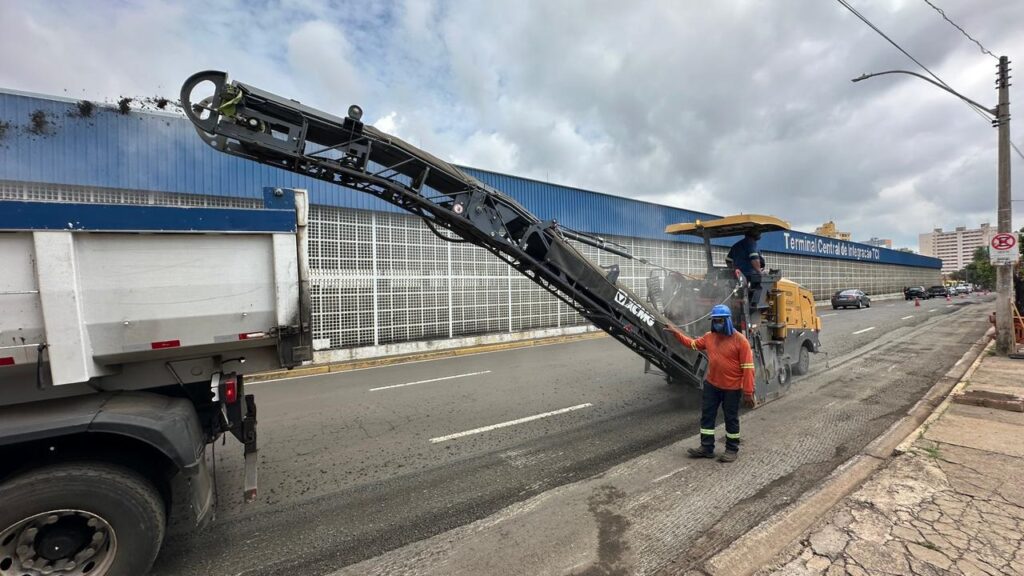 A imagem mostra uma máquina fresadora de asfalto em operação, removendo a camada superior da pavimentação de uma rua urbana. A máquina é amarela, da marca XCMG, e está posicionada atrás de um caminhão-caçamba branco e azul, para onde o material fresado está sendo transferido através de uma esteira transportadora inclinada. Ao lado da máquina, um trabalhador usa uniforme com camisa laranja, calça azul com faixas refletivas, capacete azul e máscara facial, indicando cuidados com segurança. Ao fundo, há um grande prédio azul com a inscrição "Terminal Central de Integração TCI", sugerindo que o local é uma área urbana próxima a um terminal de transporte ou integração. O céu está nublado, e a cena transmite uma atividade de manutenção ou recapeamento viário.