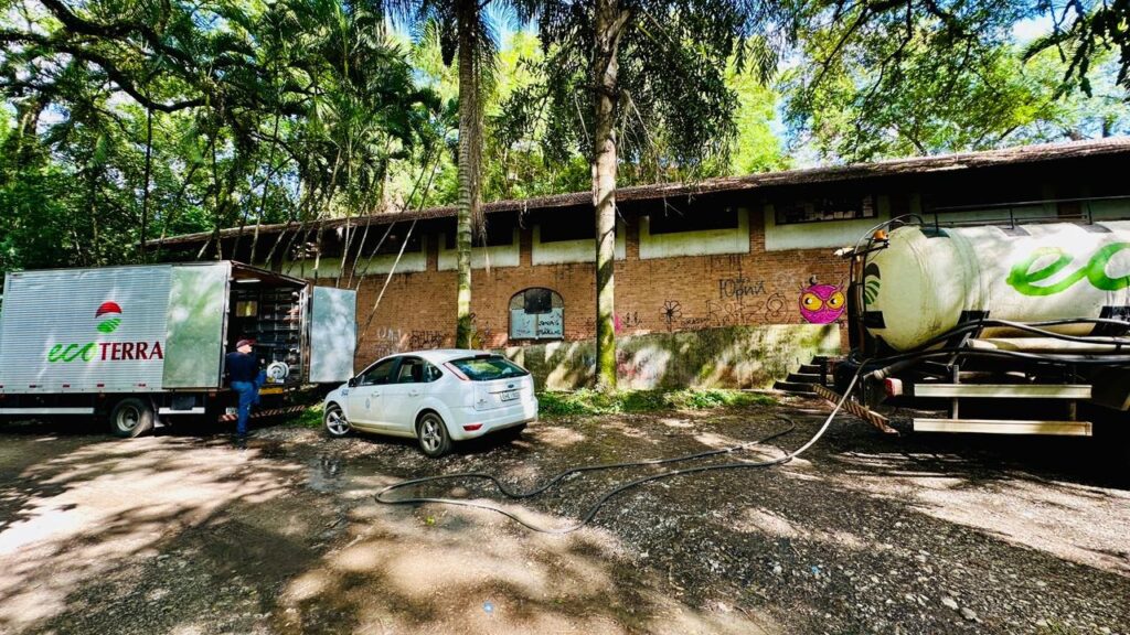 A imagem mostra uma cena industrial ou de serviço em um ambiente natural, possivelmente em uma área rural ou de floresta tropical. Há três veículos estacionados em frente a um prédio longo de tijolos com telhado de cerâmica: à esquerda, um caminhão baú branco com a porta traseira aberta e a marca "eco TERRA" em destaque; no centro, um carro branco modelo hatchback, e à direita, um caminhão-tanque branco com detalhes verdes e um tubo conectado a ele, sugerindo algum tipo de operação de transporte ou abastecimento. Ao fundo, o prédio apresenta algumas pichações, como "JONAS" e "Юрий" (em cirílico), e está cercado por árvores altas, incluindo palmeiras, que indicam um clima tropical. O chão é de terra batida, e a iluminação natural destaca as cores vibrantes das árvores e o contraste entre os veículos e o ambiente. Uma pessoa está trabalhando na parte traseira do caminhão à esquerda. No geral, a cena transmite uma atividade ligada ao meio ambiente ou serviços ecológicos, sugerida pela marca "eco TERRA", em um contexto natural e rústico.