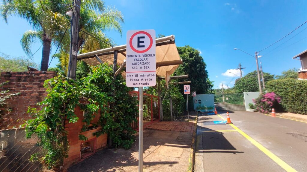 A imagem mostra uma placa de trânsito branca com um símbolo de "E" dentro de um círculo vermelho, indicando proibição de estacionamento, mas com uma exceção. Abaixo do símbolo, há um texto em português que diz: "SOMENTE VEÍCULO ESCOLAR AUTORIZADO SEG. A SEX. Por 15 minutos Pisca Alerta Acionado". Isso significa que somente veículos escolares autorizados podem estacionar ali, de segunda a sexta-feira, por no máximo 15 minutos, desde que o pisca-alerta esteja ligado. A placa está fixada em um poste metálico na calçada, ao lado de uma rua asfaltada com linhas amarelas. Ao fundo, há um muro de tijolos vermelhos, uma estrutura de madeira coberta com uma lona bege e várias árvores verdes, sugerindo um ambiente ao ar livre e provavelmente próximo a uma escola, dado o contexto da placa. Também é possível ver cones de trânsito laranja ao longo da rua e um portão ao fundo com o número 200 na parede ao lado. O céu está claro e azul, indicando um dia ensolarado. A imagem transmite uma sensação de organização e segurança no local, destinado ao embarque e desembarque de alunos.