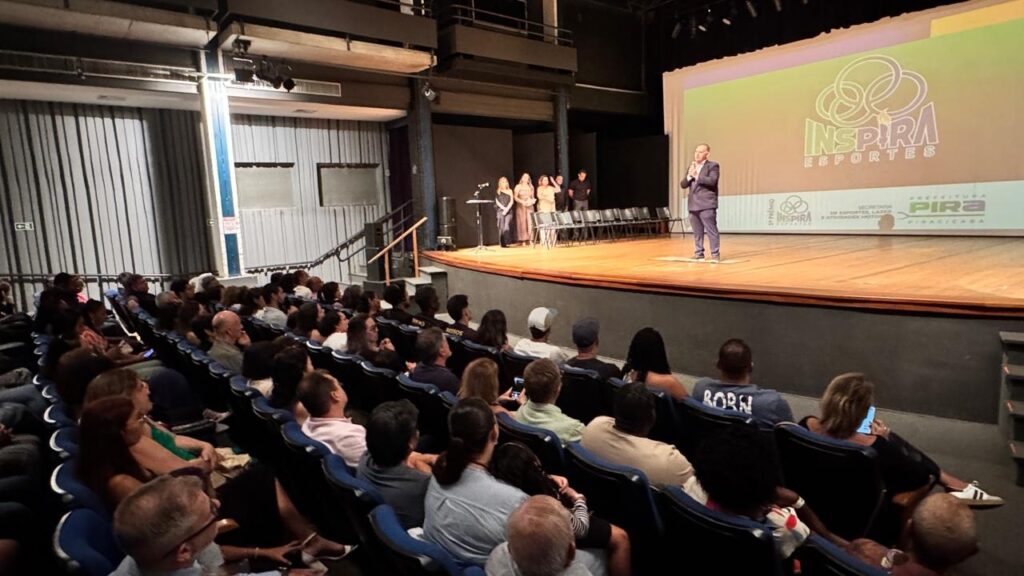 A imagem mostra um evento realizado em um auditório moderno, com arquitetura industrial, incluindo vigas de metal expostas e paredes de concreto. No palco de madeira, um homem está falando ao microfone, enquanto outras quatro pessoas (três mulheres e um homem) observam ao fundo. O palco possui várias cadeiras pretas alinhadas e uma grande tela de projeção que exibe o logotipo e o nome do evento: "INSPIRA ESPORTES" e "1º PRÊMIO INSPIRA ESPORTES", além de mencionar a Secretaria de Esportes, Lazer e Atividades Motoras e a Prefeitura de Piracicaba. A plateia está cheia, com muitas pessoas sentadas em cadeiras azuis, assistindo atentamente à apresentação. A iluminação destaca o palco, deixando o público em uma luz mais baixa, criando um ambiente focado no orador. O evento aparenta ser uma cerimônia de premiação ou uma apresentação relacionada ao esporte promovida pela prefeitura local. A foto foi tirada de um ângulo que mostra a profundidade do auditório e a quantidade de pessoas presentes.