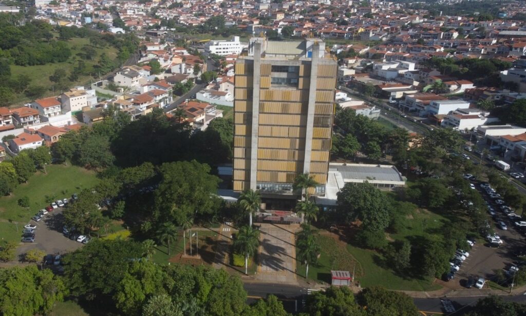 A imagem mostra uma vista aérea de um grande edifício em estilo brutalista, que parece ser um prédio público, possivelmente uma prefeitura, como sugere o texto "PREFEITURA MUNICIPAL" visível. O prédio é alto, retangular, com uma fachada marcada por painéis verticais de cor amarelada e duas colunas de concreto que sobem pela frente. Ao redor do edifício, há uma área verde com várias árvores, incluindo palmeiras, e estacionamentos com muitos carros. Próximo ao prédio principal, há uma construção menor e branca de teto plano. A região ao redor é densamente urbanizada, com muitas casas e prédios baixos, cujos telhados têm tonalidades avermelhadas. A luz do sol, provavelmente da manhã ou tarde, cria sombras longas que se projetam para o lado. O cenário mostra uma cidade extensa ao fundo, sob um céu claro e iluminado.
