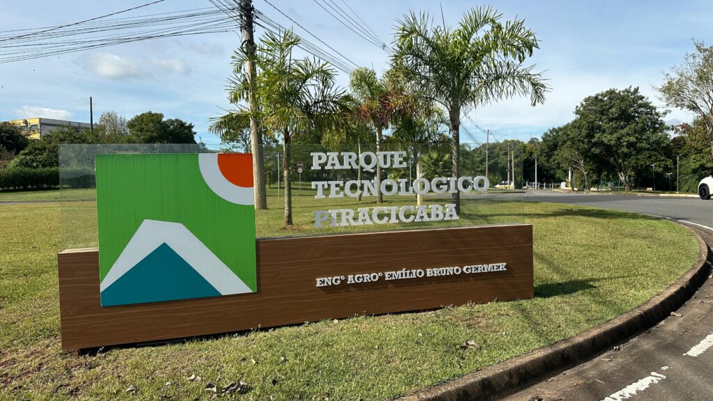 A imagem mostra uma placa grande de um parque tecnológico, chamada "Parque Tecnológico Piracicaba". A placa é composta por uma base de madeira clara com o nome "Engº Agroº Emílio Bruno Germek" em letras prateadas. Acima dessa base, há um painel de vidro transparente com o nome do parque em letras grandes e prateadas. À esquerda do texto, há um logotipo colorido que combina verde, branco, azul e laranja, formando uma figura abstrata que lembra montanhas e um círculo. A placa está situada em um gramado bem cuidado, próximo a uma estrada asfaltada. Ao fundo, vê-se várias palmeiras e vegetação verde, além de postes e fios de energia elétrica. O céu está claro, com poucas nuvens, indicando um dia ensolarado. A iluminação natural destaca as letras da placa, que refletem a luz do sol, criando um ambiente agradável e moderno, típico de um centro de inovação tecnológica.