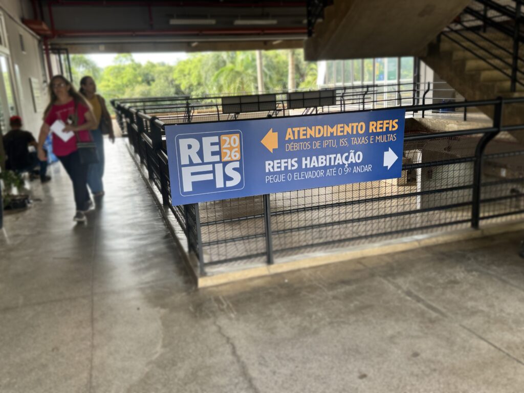 A imagem mostra o interior do prédio público, da prefeitura de Piracicaba. Em primeiro plano, há uma grade de metal preta com uma placa grande e colorida fixada nela. A placa é azul com detalhes em laranja e branco, contendo instruções sobre atendimento do programa REFIS 2026, que trata da regularização de débitos de IPTU, ISS, taxas e multas. Na placa, há duas setas: uma laranja apontando para a esquerda indicando "ATENDIMENTO REFIS DÉBITOS DE IPTU, ISS, TAXAS E MULTAS" e outra branca apontando para a direita indicando "REFIS HABITAÇÃO PEGUE O ELEVADOR ATÉ O 9º ANDAR". Ao fundo, vê-se um corredor amplo com piso de concreto polido e paredes claras. Três pessoas estão visíveis: duas mulheres caminhando, uma usando camiseta vermelha e outra amarela, e um homem sentado à esquerda. A iluminação é natural, provavelmente vinda de grandes janelas ou uma área aberta, e ao fundo é possível ver árvores verdes, sugerindo que o prédio tem uma vista para o exterior. O ambiente parece organizado e moderno, com um design funcional para atender o público que busca serviços relacionados ao REFIS.