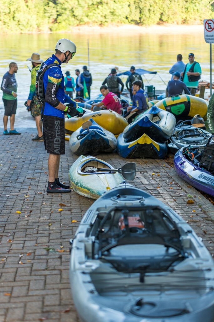 A imagem mostra um grupo de pessoas em uma rampa de acesso a um rio, preparando-se para uma atividade de caiaque. No centro da foto, um homem está em pé, vestindo um capacete branco, uma camiseta azul com a palavra "FORADO" e shorts pretos com a palavra "ADRESS". Ele está cercado por vários caiaques coloridos — um cinza em primeiro plano, amarelo, azul e roxo ao redor — e algumas pessoas sentadas ou em pé, aparentemente se preparando para entrar na água. O cenário é ao ar livre, com uma vegetação verde densa ao fundo e o rio calmo à direita da imagem. Há uma placa que indica "EMBARQUE E DESEMBARQUE", sugerindo que o local é uma área designada para iniciar ou terminar o passeio de caiaque. A luz natural do dia ilumina a cena, e todos parecem focados na atividade que estão prestes a realizar. A atmosfera transmite uma sensação de preparo e expectativa antes de uma aventura aquática em grupo.