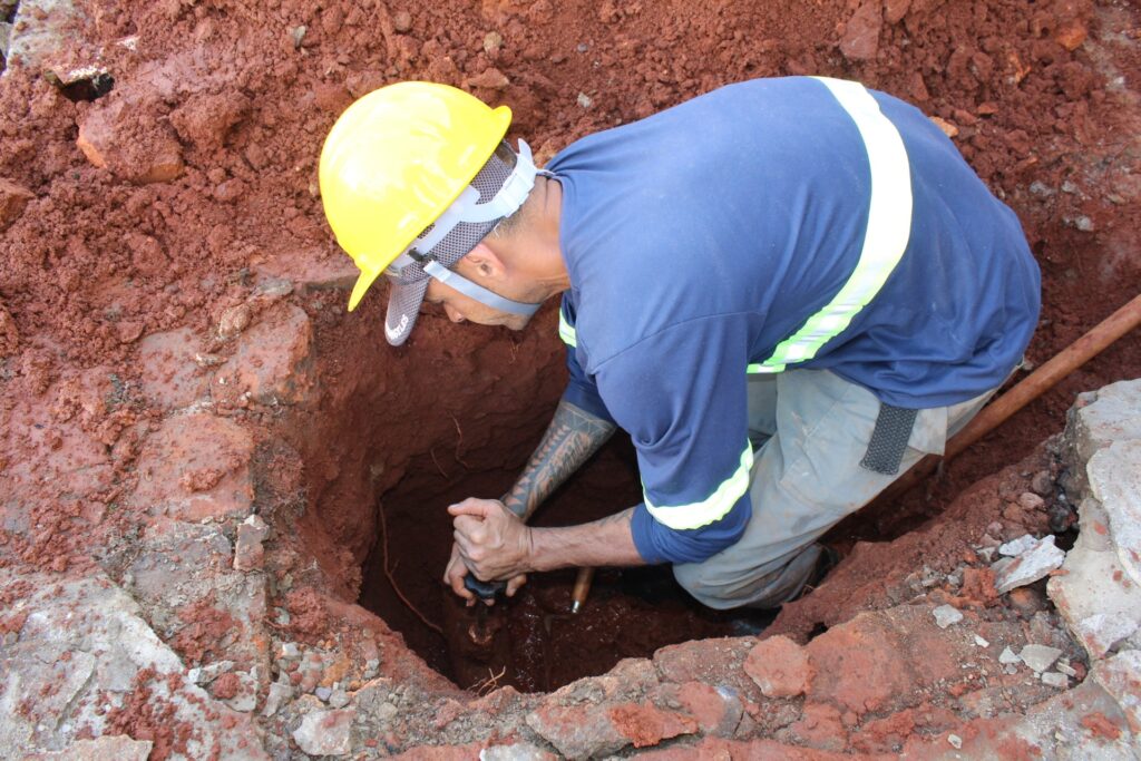 A imagem mostra um trabalhador, possivelmente de construção ou serviços públicos, dentro de uma vala profunda cavada no solo. O solo ao redor é de cor avermelhada, aparentando ser argiloso e úmido. O homem está agachado ou curvado dentro da vala, usando um capacete amarelo de segurança e uma camisa azul com faixas refletivas amarelas. Ele segura uma ferramenta, sugerindo que está realizando algum tipo de reparo ou instalação. Ao redor da vala, há pedaços de concreto quebrado, indicando que a área foi aberta para acesso a alguma infraestrutura subterrânea. A iluminação é natural, provavelmente durante o dia, e o ambiente aparenta ser uma obra ou manutenção externa.