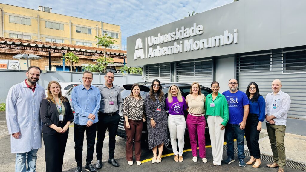 A imagem mostra um grupo de onze pessoas (seis mulheres e cinco homens) posando para uma foto em frente a um prédio com a inscrição "Universidade Anhembi Morumbi". Eles estão alinhados lado a lado, sobre um chão de asfalto, e parecem estar em um ambiente externo durante o dia. As pessoas vestem roupas variadas, desde jalecos brancos até camisetas e roupas casuais. Uma mulher no centro usa uma camiseta roxa com a palavra "Epilepsia" e um homem à direita veste uma camiseta azul com a inscrição "pilepCIA ALICE". Todos estão sorrindo e olhando para a câmera. Ao fundo, além do prédio cinza com a inscrição da universidade, há um carro escuro estacionado e uma estrutura amarela à esquerda. O clima parece ser claro com luz natural. A imagem transmite um ambiente profissional e amigável, possivelmente relacionado a uma iniciativa ou campanha sobre epilepsia.