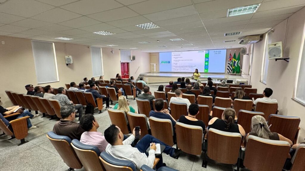 A imagem mostra uma reunião ou apresentação em um auditório. Uma mulher, vestindo uma blusa amarela, está em pé na frente, falando para um público sentado em várias fileiras de cadeiras de madeira com assentos estofados. O público é formado por cerca de 40 a 50 pessoas, que parecem estar prestando atenção à apresentação. Na frente da sala, há uma grande tela digital onde se lê "PREFEITURA PIRA", "3 - CRONOGRAMA" e a data e horário "21.02.2024 16:24". Ao lado da tela, há três bandeiras, provavelmente a bandeira do Brasil, a do estado e a do município, indicando que o evento é oficial e relacionado ao governo local. O ambiente é claro e bem iluminado, com teto rebaixado e iluminação fluorescente. As cadeiras estão dispostas em formato semicircular, voltadas para o palco, favorecendo a visualização da apresentação. A atmosfera sugere um encontro formal, possivelmente uma reunião administrativa ou uma sessão de planejamento municipal.