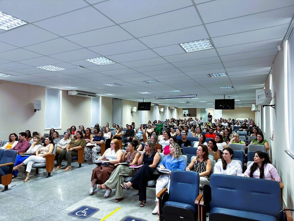 A imagem mostra um auditório grande e moderno, visto de um ângulo alto e diagonal. O espaço está cheio de pessoas sentadas em cadeiras azuis com braços de madeira, a maioria mulheres, vestidas de forma casual e casual elegante. As pessoas parecem estar atentas, possivelmente assistindo a uma palestra ou apresentação que ocorre fora do quadro, à esquerda. O teto tem luminárias retangulares embutidas que iluminam bem o ambiente. O piso é cinza com manchas, e há uma placa no canto inferior esquerdo indicando acessibilidade para cadeirantes. A atmosfera geral transmite um ambiente profissional e educacional, com um público engajado.