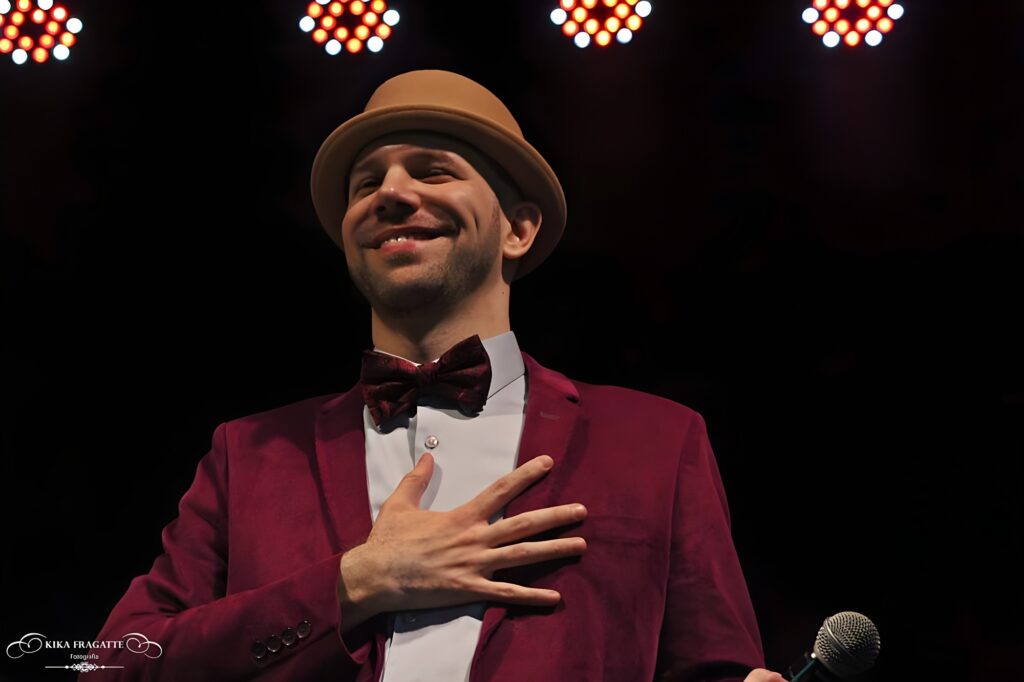 A imagem mostra um homem em um palco, fotografado de um ângulo baixo, o que dá uma sensação de imponência. Ele veste um chapéu estilo fedora bege, um blazer de veludo vinho, uma camisa branca e uma gravata borboleta vermelha com padrão. O homem sorri amplamente, demonstrando alegria, e tem a mão direita sobre o peito, gesto que pode indicar gratidão ou emoção. Ele tem barba curta e pele clara. O fundo é escuro, destacando o sujeito, e há quatro luzes circulares acesas acima dele, com tons alaranjados e brancos que criam um efeito dramático. No canto inferior direito, há um microfone prateado parcialmente visível, sugerindo que ele está em uma apresentação. No canto inferior esquerdo, há uma marca d'água com o texto "KIKA FRAGATTE Fotografia". A cena transmite a imagem de um artista recebendo aplausos ou agradecendo ao público após uma performance.