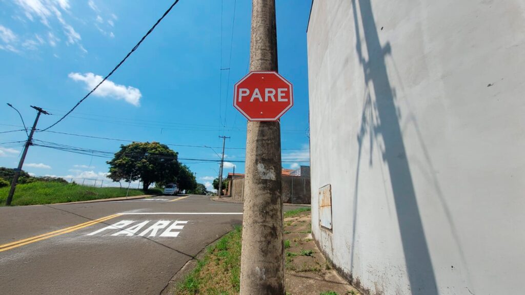 A imagem mostra uma placa de trânsito vermelha em formato octogonal com a palavra "PARE" escrita em branco, fixada em um poste de concreto. A placa está localizada em uma esquina de rua, onde o chão também tem a palavra "PARE" pintada em branco sobre o asfalto. Ao fundo, há uma parede branca e uma árvore verde, além de fios elétricos visíveis no céu azul claro. A cena ocorre em um dia ensolarado, com luz forte que cria sombras nítidas. A palavra "PARE" indica que provavelmente a imagem foi tirada em um país de língua portuguesa, como o Brasil.