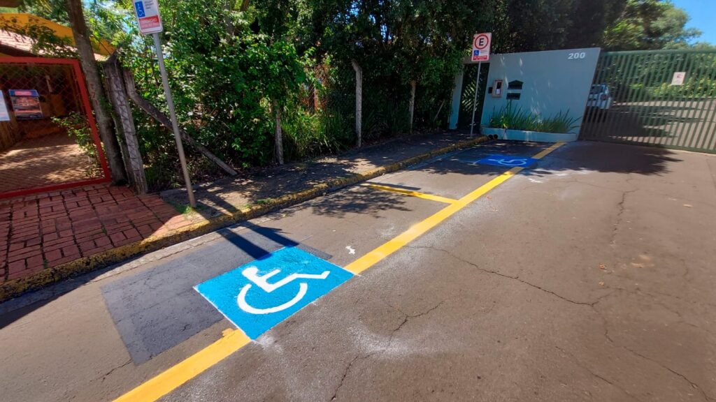 A imagem mostra duas vagas de estacionamento reservadas para pessoas com deficiência, identificadas pelo símbolo internacional de acessibilidade (cadeirante) pintado no chão em azul com o símbolo branco. As vagas são delimitadas por linhas amarelas no chão e estão localizadas em frente a um portão azul claro, que parece ser a entrada de um imóvel residencial ou comercial. Acima de cada vaga, há uma placa com a letra "E" dentro de um círculo vermelho, indicando estacionamento, e o símbolo de acessibilidade. Em uma das placas está escrito "USO OBRIGATÓRIO DE CARTÃO", indicando que para estacionar ali é necessário possuir o cartão de estacionamento especial para pessoas com deficiência. À esquerda, há uma calçada com um portão vermelho e algumas árvores que projetam sombras no chão. O número "200" está pintado na parede azul clara, provavelmente indicando o endereço do local. A iluminação é forte, típica de um dia ensolarado.