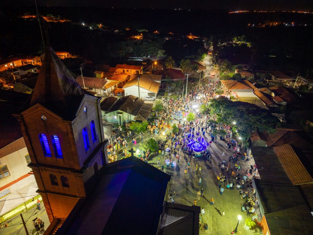 A imagem mostra uma vista aérea noturna de uma praça movimentada em uma pequena cidade. Em primeiro plano, à esquerda, há uma igreja com uma torre alta e uma cruz no topo, iluminada por luzes azuis que destacam suas janelas. A praça está cheia de pessoas, algumas caminhando, outras em grupos ou sentadas em mesas, sugerindo um evento ou festival acontecendo. No centro da praça, há uma fonte circular iluminada por luzes azuis, que contrasta com a iluminação amarela quente das lâmpadas de rua e das casas ao redor, que possuem telhados de cerâmica laranja. Ao fundo, é possível ver colinas com algumas luzes dispersas, indicando áreas residenciais ou rurais. A atmosfera é vibrante e acolhedora, típica de uma celebração comunitária noturna.