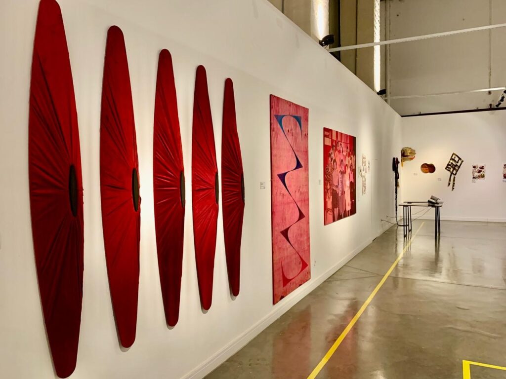 A imagem mostra o interior de uma galeria de arte moderna, com paredes brancas e piso de concreto polido. Na parede à esquerda, há cinco esculturas verticais vermelhas, alongadas e com um formato que lembra olhos ou formas orgânicas, cada uma com uma fenda central. Ao lado dessas esculturas, há uma grande pintura abstrata em tons de rosa, azul e vermelho, com formas onduladas. Mais à direita, está pendurada uma fotografia em tons vermelhos que retrata um grupo de pessoas. No fundo da galeria, é possível ver uma mesa pequena com alguns objetos sobre ela e outras obras menores penduradas na parede. A iluminação é forte e vem do teto, criando um ambiente claro e contemporâneo. O espaço transmite uma sensação de modernidade e organização, com linhas que guiam o olhar para o fundo do ambiente.