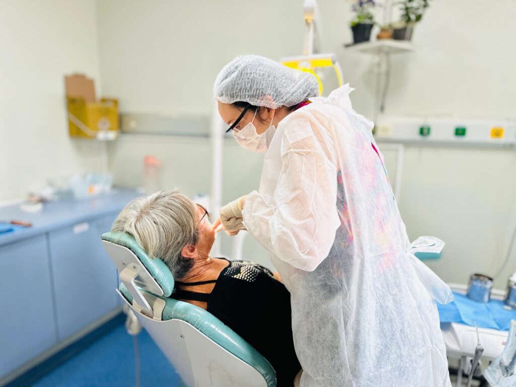 A imagem mostra um ambiente clínico, provavelmente um consultório odontológico ou médico. Uma profissional de saúde, vestindo equipamento de proteção individual (EPI) completo — incluindo touca, máscara facial, óculos e avental descartável — está atendendo uma paciente. A profissional parece estar examinando ou realizando um procedimento na boca da paciente, que está reclinada em uma cadeira odontológica de cor verde água. A paciente é uma mulher mais velha, com cabelo grisalho curto e óculos, vestindo uma blusa preta com detalhes brancos. O ambiente é limpo e bem iluminado, com paredes brancas, armário azul claro ao fundo, pequenas plantas em uma prateleira e equipamentos médicos na parede, como tomadas para gases medicinais e uma caixa amarela para descarte de materiais perfurocortantes. A cena transmite um clima de cuidado e profissionalismo.