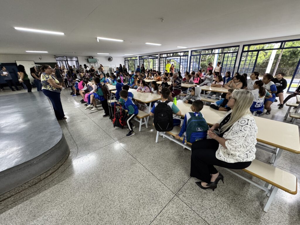 A imagem mostra um ambiente interno, provavelmente uma sala de múltiplo uso ou refeitório de uma escola, onde várias crianças e alguns adultos estão reunidos para uma reunião ou evento escolar. As crianças estão sentadas em longas mesas com bancos fixos, muitas vestindo uniformes escolares que incluem camisas azuis e brancas com a palavra "Piracicaba" e logos relacionados à educação, sugerindo que a escola está localizada em Piracicaba, Brasil. À esquerda da imagem, há um pequeno palco elevado onde alguns adultos estão em pé, aparentemente falando ou se preparando para falar com os alunos. No centro e à direita, as crianças estão sentadas, algumas com mochilas coloridas ao lado. Também há adultos sentados entre as crianças, possivelmente professores ou responsáveis. A iluminação é feita por lâmpadas fluorescentes no teto, e grandes janelas laterais permitem a entrada de luz natural, mostrando árvores do lado externo. O chão é de azulejos cinza com um padrão. Em primeiro plano, uma mulher com uma blusa branca de renda e calça preta observa a cena. O ambiente transmite uma atmosfera de organização e atenção, típica de um momento formal de orientação ou reunião escolar, possivelmente no início do ano letivo.