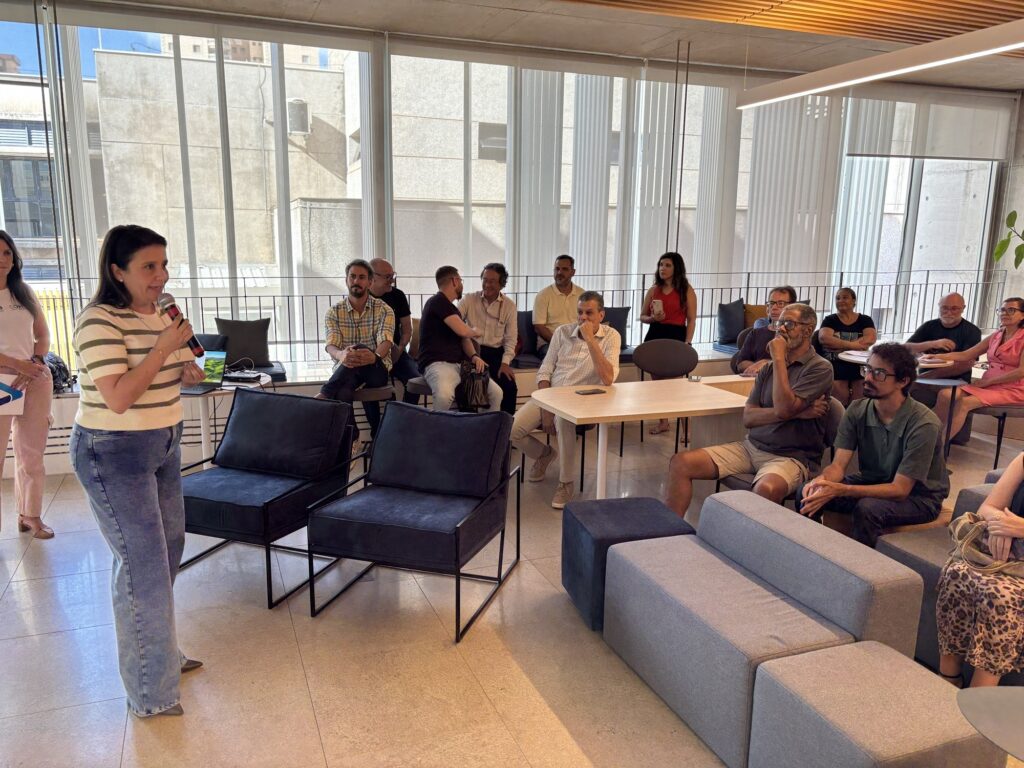 A imagem mostra uma mulher falando em um microfone para um grupo de pessoas sentadas em um ambiente moderno e bem iluminado. Ela está usando uma blusa listrada e jeans, e parece estar fazendo uma apresentação ou conduzindo uma reunião. O local tem janelas grandes que deixam entrar bastante luz natural, e há cadeiras e sofás distribuídos pelo espaço, que é decorado de forma simples e contemporânea. As pessoas na audiência estão atentas, algumas sentadas em cadeiras pretas e outras em sofás cinza. Ao fundo, é possível ver prédios urbanos através das janelas, indicando que o ambiente está em uma área urbana. A atmosfera parece ser profissional e colaborativa.