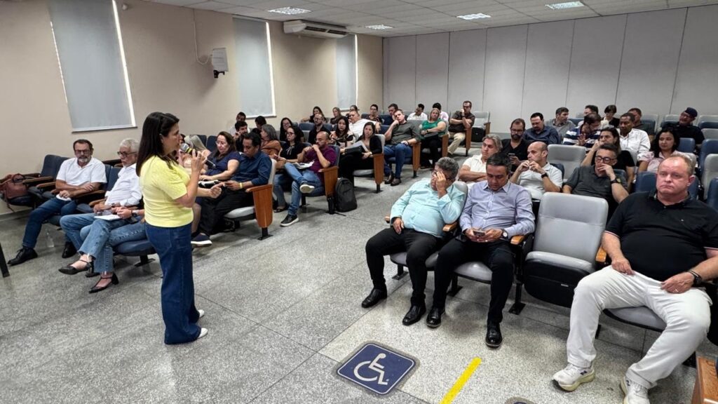 A imagem mostra uma mulher palestrando em uma sala de conferências ou auditório. Ela está à esquerda, segurando um microfone e vestindo uma camiseta amarela e calças jeans largas. À sua frente, um grupo de pessoas está sentado em cadeiras organizadas em fileiras, prestando atenção à apresentação. O público é composto por adultos, tanto homens quanto mulheres, vestidos de forma casual ou semi-formal. A sala tem paredes claras, um piso cinza e iluminação fluorescente no teto. Há janelas à esquerda com cortinas fechadas. No chão, próximo ao primeiro plano da imagem, há um símbolo de acessibilidade para cadeirantes, indicando que o local é adaptado. A atmosfera parece ser de concentração e interesse na palestra.