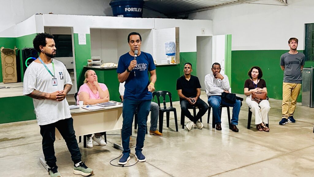 A imagem mostra uma reunião ou encontro em um ambiente interno, possivelmente uma sala de aula ou auditório simples com paredes pintadas de branco e verde. No centro, um homem está de pé, falando ao microfone, vestindo uma camiseta azul com a inscrição "ante & SEM PARAR" e uma referência bíblica "JOÃO 4:14". Ao lado dele, outro homem também está em pé, vestindo uma camiseta branca larga e calça preta. Sentados em cadeiras de plástico pretas, há quatro adultos: um homem de camiseta preta, um homem de camisa branca e jeans, uma mulher de blusa marrom e calça branca, e um homem de camiseta cinza da marca Puma e calça bege, que está em pé. Ao fundo, há uma mesa com uma mulher sentada, que parece estar observando ou tomando notas. Também é possível ver um tanque d’água azul da marca "FORTLEV", alto, colocado sobre uma estrutura. Há equipamentos eletrônicos, como caixas de som, e um ambiente simples e funcional, com piso de cimento polido. A cena sugere um momento de fala ou apresentação em um evento comunitário, religioso ou educacional.