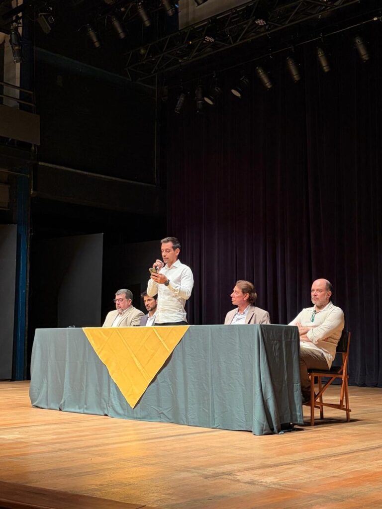 A imagem mostra cinco homens em um palco de madeira, provavelmente em um teatro ou auditório. Quatro deles estão sentados atrás de uma mesa longa coberta por uma toalha escura com um caminho de mesa amarelo em forma de losango no centro. O quinto homem está em pé, ao centro-esquerda da mesa, vestindo uma camisa branca e olhando para um celular que segura com as duas mãos, parecendo estar falando ou lendo algo do aparelho. À esquerda do homem em pé, há dois homens sentados; um deles usa um blazer bege claro e óculos, e o outro está parcialmente oculto. À direita do homem em pé, há mais dois homens sentados, um deles usando um blazer cor de areia e olhando para o homem em pé, e o outro, à extrema direita, calvo e com barba, veste uma camisa clara e calças cáqui, está com os braços cruzados e olhando para a câmera. Ao fundo, há uma cortina escura, possivelmente roxa ou preta, e acima deles, um suporte com várias luzes de palco. A iluminação é quente e direcionada, destacando o grupo no palco e criando sombras no chão de madeira. A cena sugere um evento formal, como uma mesa redonda, uma conferência ou uma apresentação.
