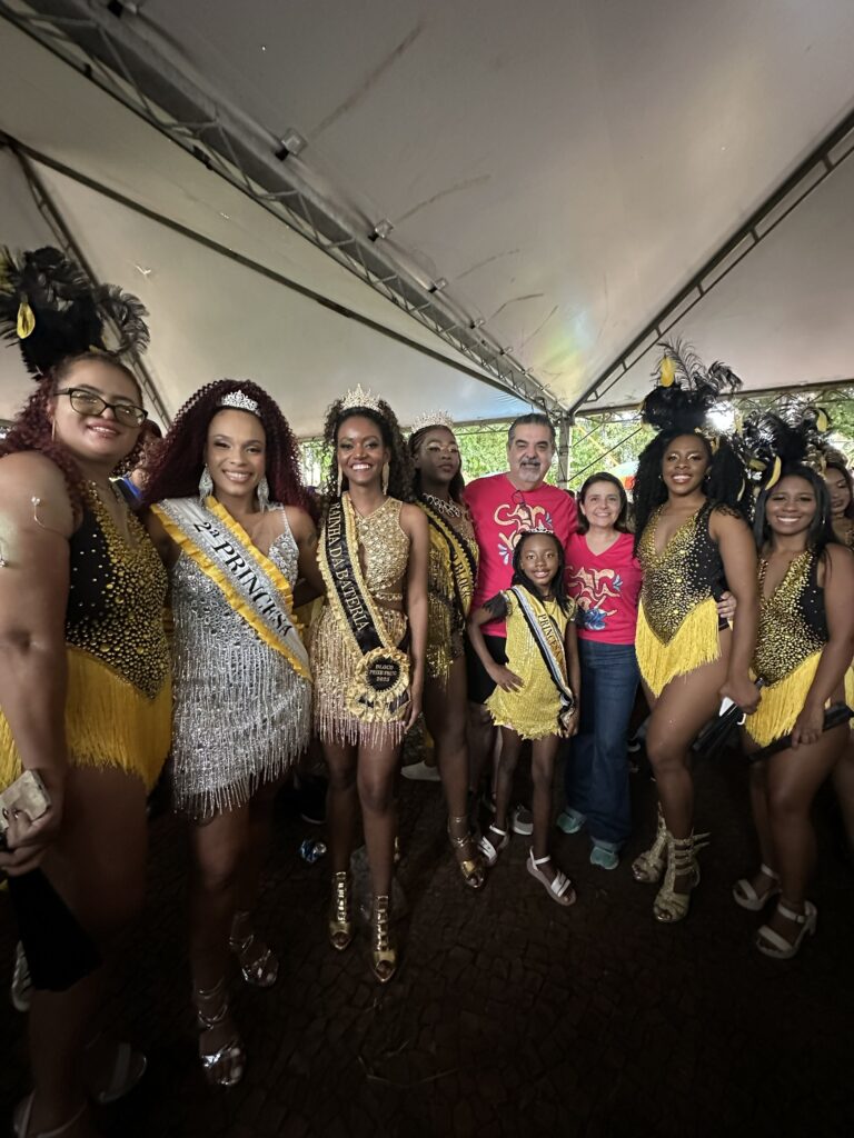 A imagem mostra um grupo de nove pessoas posando sob uma tenda branca à noite. Na frente, há cinco mulheres vestidas com trajes carnavalescos elaborados, cheios de lantejoulas, franjas e penas, em cores como dourado, prata e preto. Elas usam faixas com títulos como "2ª PRINCESA", "RAINHA DA BATERIA" e "PRINCESA", indicando seus papéis em um bloco de Carnaval chamado "BLOCO PEIXE FRITO 2023". Uma menina pequena também está vestida com um traje dourado semelhante, entre as mulheres. Ao fundo, aparecem duas pessoas vestidas com camisetas vermelhas, provavelmente integrantes ou organizadores do evento. O ambiente é iluminado artificialmente, destacando o brilho dos trajes. O chão é de pedra escura, e o clima geral é de celebração e festa.