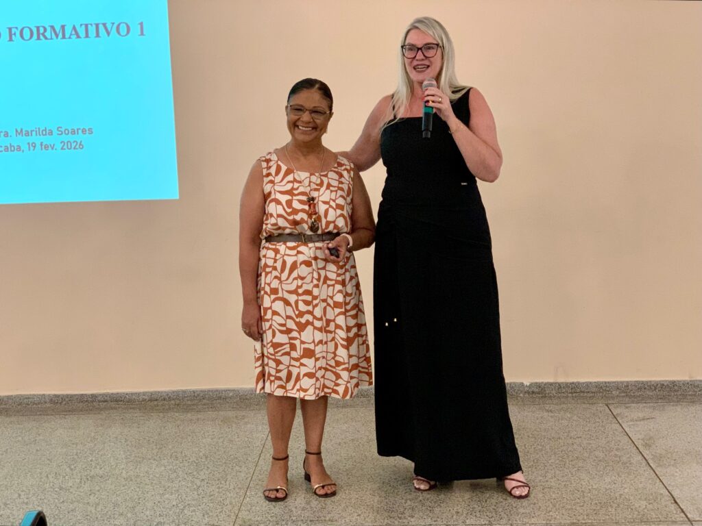 A imagem mostra duas mulheres em um ambiente interno, possivelmente uma sala de aula ou auditório, durante um evento ou apresentação. A mulher à esquerda tem cabelo escuro preso em um coque, usa óculos e um vestido laranja com estampa branca, acompanhado de um cinto marrom e sandálias. Ela segura um controle remoto, provavelmente para a apresentação. A mulher à direita é loira, também usa óculos e um vestido preto longo, além de sandálias. Ela está falando ao microfone e apoia a mão no ombro da outra mulher, transmitindo uma sensação de apoio ou parceria. Atrás delas, há uma parede clara e, à esquerda da imagem, uma tela de projeção azul com o texto "FORMATIVO 1", "ra. Marilda Soares" e "caba, 19 fev. 2026", indicando que se trata de um evento formativo realizado em 19 de fevereiro de 2026. A atmosfera parece ser amigável e profissional, com as mulheres sorrindo e interagindo de forma positiva.
