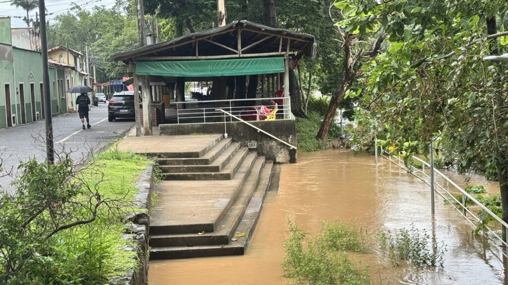 A imagem mostra uma área urbana ou semi-rural afetada por uma enchente. A água barrenta de um rio ou canal transbordou, invadindo escadas de concreto e uma calçada. À direita, há árvores verdes e densas que margeiam o curso d’água. No centro, as escadas estão parcialmente submersas, com um corrimão branco ao lado. À esquerda, uma pessoa caminha segurando um guarda-chuva preto, indicando que provavelmente está chovendo ou que choveu recentemente. Mais ao fundo, há uma estrutura de madeira com um toldo verde, onde algumas pessoas estão sentadas. Também é possível ver um carro preto estacionado na rua elevada, ao lado dessa estrutura. O céu parece nublado, dando uma atmosfera cinzenta e úmida à cena. A imagem transmite a sensação de um dia chuvoso e a força da natureza ao invadir áreas construídas pelo homem.
