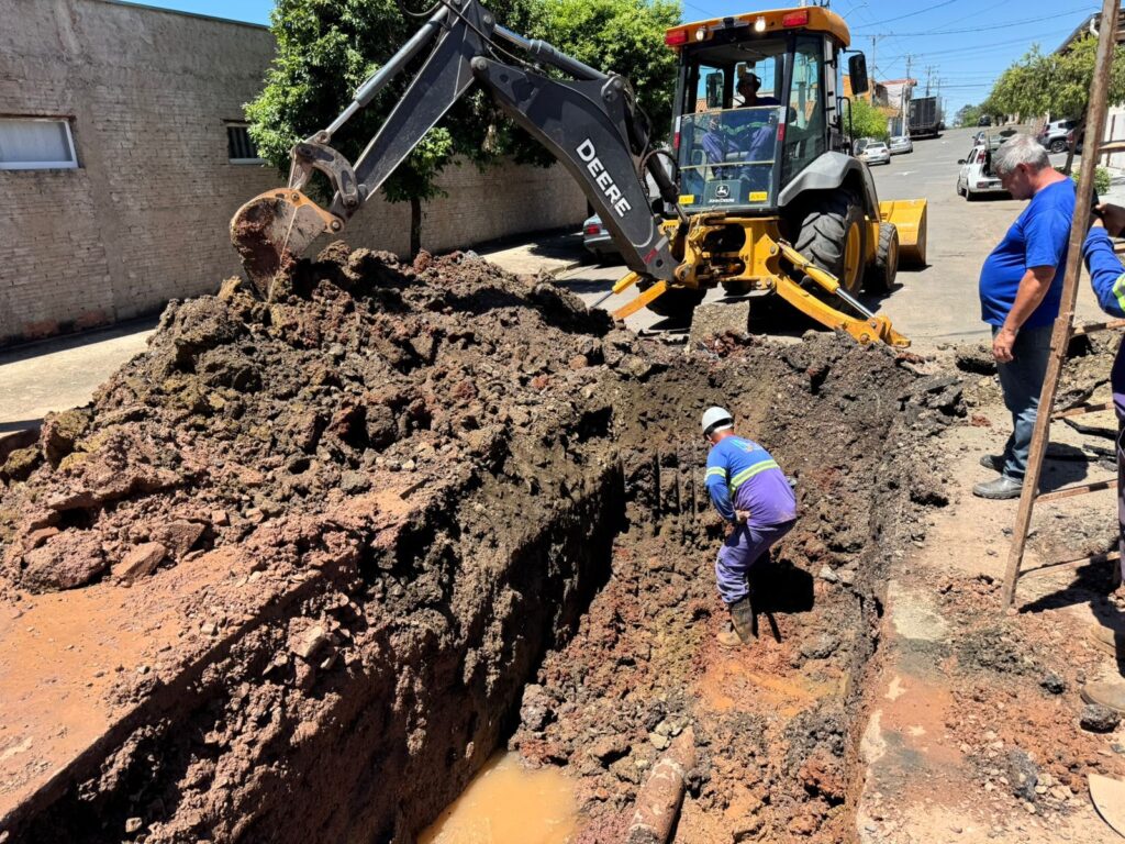 A imagem mostra uma escavadeira retroescavadeira da marca John Deere trabalhando em uma rua residencial durante o dia. A máquina está escavando uma vala profunda, onde há um pouco de água acumulada no fundo. Quatro homens estão presentes: um operador dentro da cabine da máquina, um trabalhador com capacete branco e uniforme azul dentro da vala, outro homem de camiseta azul observando a operação na calçada, e uma parte de outra pessoa à direita da imagem. Há uma escada de madeira apoiada na lateral da vala para acesso. Ao redor, é possível ver uma parede de tijolos, árvores, carros estacionados e casas ao fundo, tudo sob um céu azul claro. A cena sugere um trabalho de manutenção ou instalação de tubulação, possivelmente de água ou esgoto.