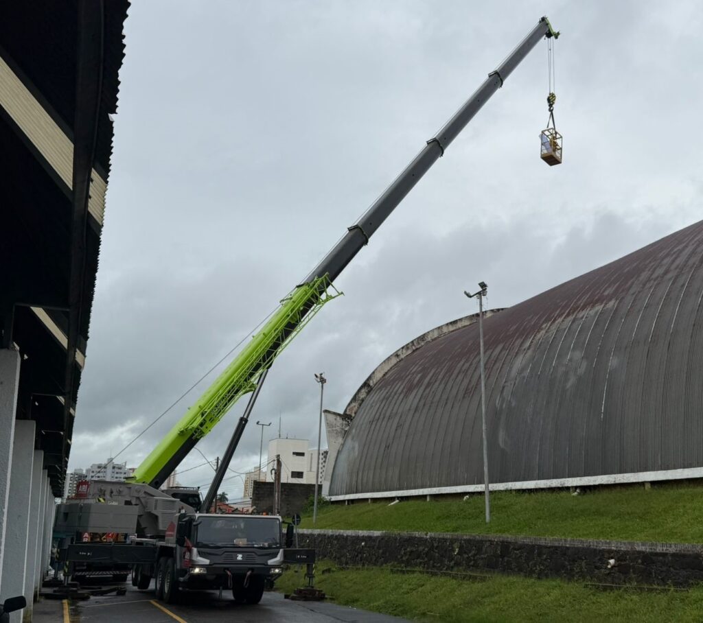 A imagem mostra um guindaste móvel grande, posicionado em uma rua estreita entre um prédio à esquerda e uma grande estrutura curva à direita, que parece ser o telhado de um galpão ou armazém. O guindaste tem um braço telescópico longo e levantado diagonalmente para cima, com a base em verde-limão e a parte superior em cinza escuro. Preso ao gancho do guindaste, há uma cesta amarela para pessoas, provavelmente usada para manutenção ou inspeção em altura. O céu está nublado, com tons de cinza, sugerindo um dia chuvoso ou nublado. Ao fundo, vê-se uma área com grama e um muro de pedras. O guindaste está identificado com as letras "ARI" na lateral, e o caminhão que o transporta tem o logo "Z" na frente, possivelmente da marca Zoomlion. A foto foi tirada de um ângulo baixo, destacando a altura e o alcance do braço do guindaste.