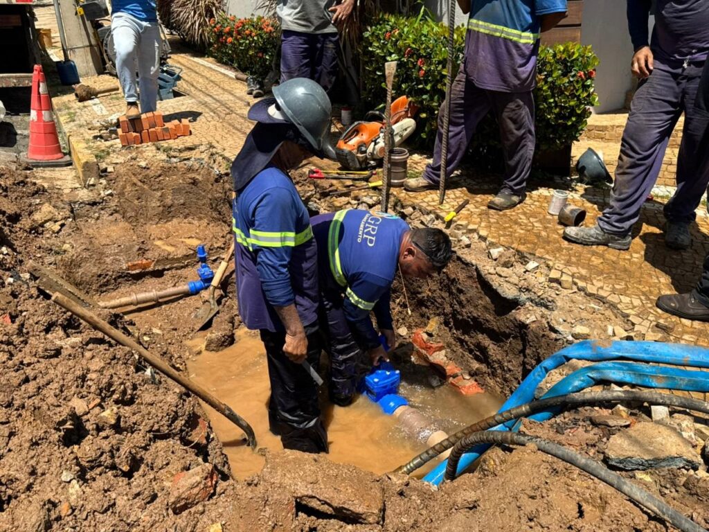 A imagem mostra uma equipe de trabalhadores realizando reparos em uma rede de água em uma área urbana. Dois trabalhadores estão dentro de uma vala cheia de água lamacenta, manipulando uma válvula azul conectada a um grande cano. Eles vestem uniformes azuis com faixas refletivas amarelas e capacetes pretos, sendo que um dos uniformes tem a sigla "GRP" e a palavra "SANEAMENTO". Ao redor da vala, outros trabalhadores observam ou auxiliam. O chão ao redor é de terra com pedaços de pavimento e pedras, e há várias ferramentas espalhadas, como serra, pás, e mangueiras azuis. Um cone de sinalização vermelho e branco está posicionado próximo, indicando que o local está em manutenção. A cena ocorre em um dia ensolarado, com luz natural forte, e retrata o trabalho manual necessário para manutenção da infraestrutura de saneamento.