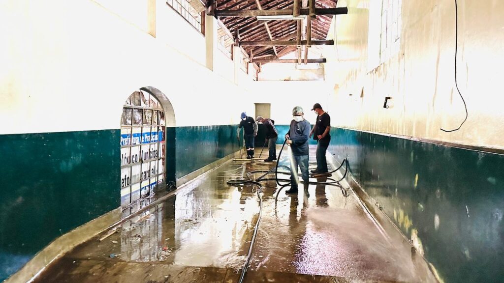 A imagem mostra um corredor interno longo e estreito, com paredes pintadas em dois tons: a parte superior em tom claro e a inferior em verde escuro. O chão está molhado e reflete a luz, indicando que está sendo limpo. Quatro homens, todos usando máscaras faciais e roupas de trabalho simples, estão envolvidos na limpeza do local. Um deles, mais próximo da câmera, segura uma mangueira preta, enquanto os outros três, mais ao fundo, utilizam rodos para espalhar a água pelo chão. O teto é alto, com estrutura de madeira aparente, e há uma janela arqueada à esquerda, pela qual entra luz natural. Na janela, há um cartaz escrito em português com as palavras "Prefeitura de Pira...", "Módulo Municipal" e "Reforma Geral", sugerindo que o local está passando por uma reforma geral organizada pela prefeitura municipal de uma cidade chamada Pira (provavelmente Piraí ou Piracicaba, por exemplo). A perspectiva da foto cria uma sensação de profundidade, com os trabalhadores distribuídos ao longo do corredor, e a iluminação é uma mistura de luz natural e artificial. A cena transmite a ideia de um ambiente institucional em processo de manutenção ou renovação durante o período da pandemia, dada a presença das máscaras.