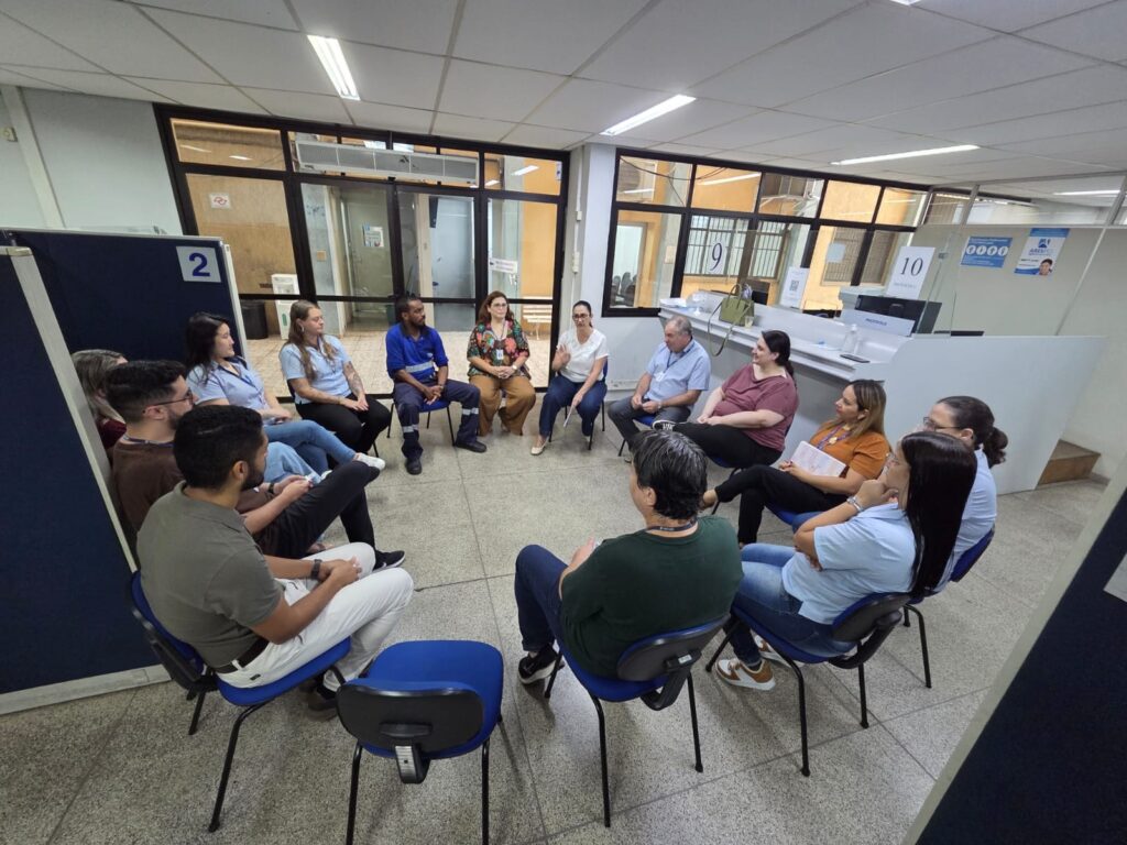 A imagem mostra um grupo de pessoas sentadas em círculo, em cadeiras azuis, dentro de um ambiente de escritório. São cerca de treze pessoas, incluindo homens e mulheres, com idades variadas. Algumas mulheres vestem camisetas polo azuis claras, enquanto um homem usa um uniforme azul. Uma mulher em pé, vestindo camisa branca e calça escura, parece estar falando ou conduzindo a reunião. O ambiente é iluminado por luzes fluorescentes no teto, com divisórias de vidro ao fundo identificadas pelos números 9 e 10, além de uma divisória azul marcada com o número 2. Ao fundo, há um balcão com a placa "PROTOCOLO". O chão é de cor cinza com aspecto granuloso. A cena transmite um clima profissional e colaborativo, possivelmente uma reunião de trabalho ou treinamento.
