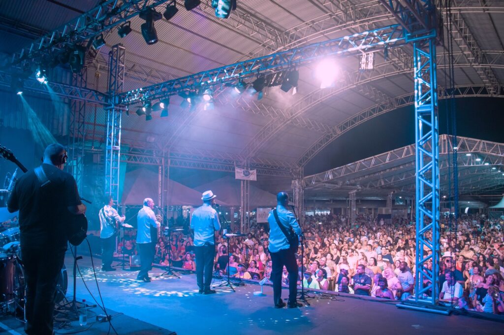 A imagem mostra um show ao vivo em um grande palco coberto por uma estrutura metálica. Cinco músicos estão se apresentando; quatro deles vestem camisas claras e calças escuras, e um deles usa um chapéu branco. Outro músico, com camisa preta, toca baixo na frente. O palco é iluminado por luzes coloridas em tons de azul e roxo, criando um ambiente vibrante. À frente do palco, uma grande plateia bastante animada observa a apresentação, com muitas pessoas sorrindo e vestindo roupas casuais, algumas até com camisas de times de futebol. O local parece ser ao ar livre ou semiaberto, com um teto de metal ondulado e várias estruturas de suporte visíveis. A atmosfera geral é de festa e celebração, provavelmente em um evento cultural ou festival. Ao fundo, é possível ver uma faixa com a palavra "IMPERIAL".