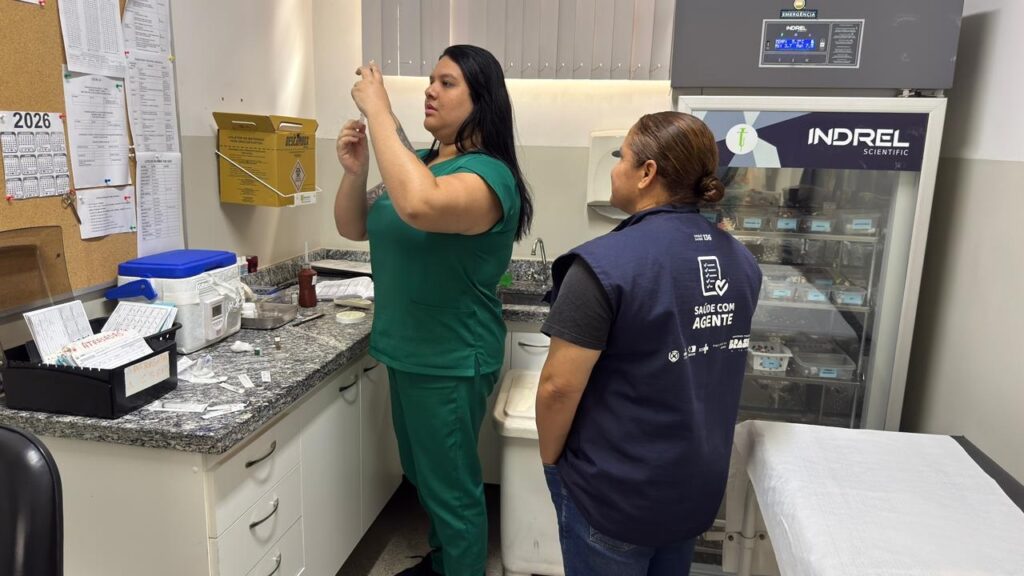 A imagem mostra um ambiente clínico, possivelmente uma sala de vacinação ou um posto de saúde. No centro, há uma profissional de saúde vestindo um uniforme verde, preparando uma seringa com uma vacina ou medicamento. Ao lado dela, uma outra mulher, vestindo um colete azul com a inscrição "SAÚDE COM AGENTE", observa atentamente o procedimento. Ao fundo, há um armário refrigerado da marca "INDREL SCIENTIFIC", provavelmente usado para armazenar vacinas ou medicamentos que precisam ser mantidos em baixa temperatura, indicada no visor em 5,2°C. Na parede, um recipiente amarelo para descarte de materiais perfurocortantes está fixado, com a palavra "DESCARPACK". Também é possível ver um quadro de avisos com calendários e documentos, e uma mesa com papéis e outros materiais de trabalho. A presença do logo do SUS e do governo do Brasil sugere que o local é uma unidade pública de saúde. A imagem transmite um ambiente organizado, limpo e focado na imunização ou cuidados médicos.
