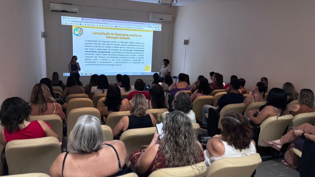 A imagem mostra uma sala de aula ou auditório onde está acontecendo uma apresentação ou palestra voltada para educadores. Na frente da sala, uma mulher está falando e apontando para um grande telão onde está projetada uma apresentação em português intitulada "Apropriação da linguagem escrita na Educação Infantil". O texto da apresentação fala sobre a importância de um processo educativo estruturado para que as crianças ampliem seu conhecimento e vivência da cultura escrita, destacando o papel dos educadores em proporcionar contato frequente com diferentes textos e em estimular a curiosidade e o aprendizado. A plateia é composta por várias pessoas sentadas em cadeiras dispostas em fileiras, a maioria mulheres, que estão prestando atenção à palestra. A sala é iluminada artificialmente e parece ser um ambiente dedicado a eventos educacionais ou formações. O cenário transmite uma atmosfera de aprendizado e troca de conhecimentos sobre educação infantil.