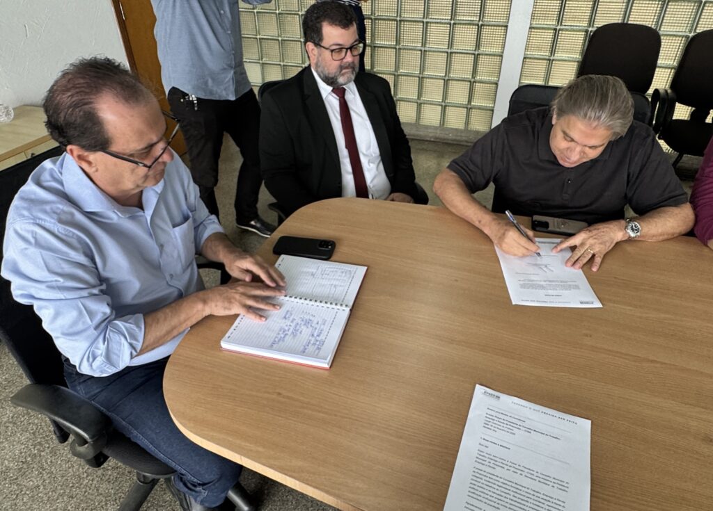 A imagem mostra uma reunião formal em um ambiente de escritório, onde três homens estão sentados ao redor de uma mesa oval de madeira clara. O homem à direita está assinando um documento, enquanto os outros dois observam. O homem do centro veste um terno preto com camisa branca e gravata vermelha, transmitindo uma imagem formal e profissional. O homem à esquerda usa uma camisa azul clara e óculos, aparentemente tomando notas em um caderno espiral. Sobre a mesa há documentos, dois celulares e uma caneta. Ao fundo, é possível ver cadeiras de escritório pretas e uma parede de blocos de vidro, que deixa o ambiente bem iluminado. Na mesa, há um documento com o cabeçalho “PIRA FAZENDO O QUE PRECISA SER FEITO” e o título “Posse do Presidente do Conselho Municipal do Trabalho, Emprego e Renda de Piracicaba - CTER”, indicando que o encontro está relacionado a uma cerimônia oficial de posse ligada à Prefeitura de Piracicaba. A atmosfera é de formalidade e trabalho administrativo, sugerindo um momento importante para a gestão pública local.
