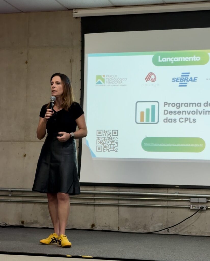 A imagem mostra uma mulher apresentando em um palco, em frente a uma grande tela de projeção. Ela está falando ao microfone e gesticulando com a mão esquerda. A mulher usa uma blusa preta de mangas curtas, saia preta de material que parece couro e tênis amarelos com detalhes pretos da marca Onitsuka Tiger. O fundo é composto por uma parede de concreto à esquerda e a tela branca de projeção à direita. Na tela, há um slide com o título "Lançamento" e os logos do Parque Tecnológico Piracicaba, Pecege e Sebrae. Também aparece um gráfico de barras e o texto "Programa de Desenvolvim das CPLs", além de um QR code e um link para o site do Parque Tecnológico de Piracicaba. O ambiente parece ser uma sala de eventos ou conferências