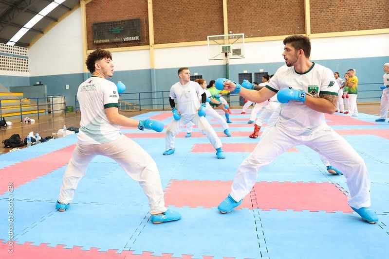 A imagem mostra um ambiente interno de um ginásio esportivo, onde um grupo de atletas está treinando karatê. Em primeiro plano, dois homens jovens estão frente a frente, em posição de combate, usando protetores azuis nas mãos e nos pés. Eles vestem camisetas brancas com detalhes verdes e calças brancas, que fazem parte do uniforme da equipe "Karate do Brasil". Ao fundo, outros atletas também praticam movimentos de karatê sobre um piso de tatames interligados nas cores azul e vermelho. O ginásio é bem iluminado, com luz natural entrando por janelas no teto. Na parede de fundo, há uma tabela de pontuação com as palavras "PIRACICABA" e "VISITANTE", indicando que o local pode ser uma arena para competições ou treinos oficiais. Também é possível ver uma cesta de basquete, evidenciando que o espaço é multiuso. No uniforme dos atletas, há o logo da Confederação Brasileira de Karatê (CBK), reforçando que eles pertencem à seleção nacional. A imagem transmite uma atmosfera de concentração, disciplina e preparação esportiva.