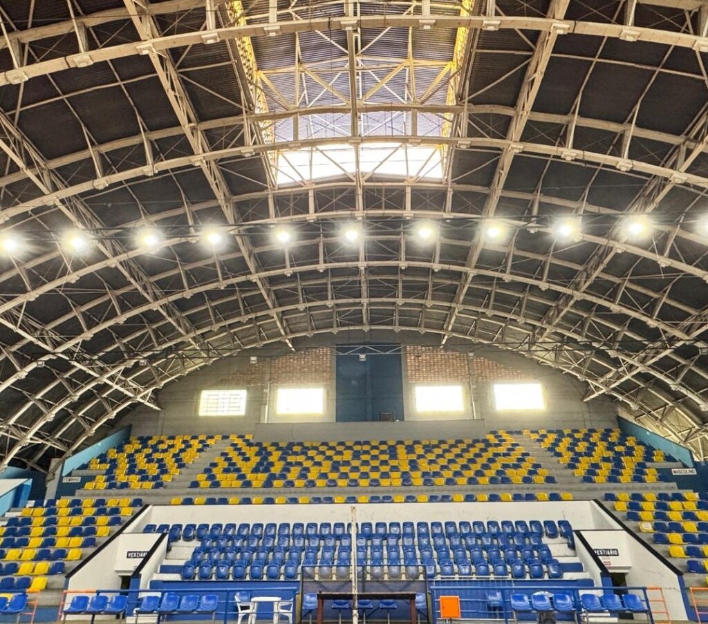 A imagem mostra o interior de um ginásio esportivo coberto, com uma estrutura de teto arqueada e robusta, feita de metal e concreto. As arquibancadas são organizadas em degraus e possuem assentos azuis e amarelos, dispostos em faixas horizontais. No centro inferior da imagem, é possível ver uma rede de vôlei, indicando que o local é utilizado para esportes indoor. Acima das arquibancadas, há janelas que permitem a entrada de luz natural, além de refletores que iluminam o espaço. Na parte inferior das arquibancadas, há portas com placas indicando “FEMININO” e “MASCULINO”, provavelmente os vestiários, já que também há uma placa com a palavra “VESTIÁRIO”. O ambiente está vazio, sem pessoas, e a iluminação natural destaca a estrutura imponente do teto e as cores vibrantes dos assentos. O ginásio aparenta ser espaçoso e bem cuidado, com uma arquitetura funcional para eventos esportivos e possivelmente outras atividades. A presença das palavras em português sugere que o local fica em um país lusófono, como o Brasil.